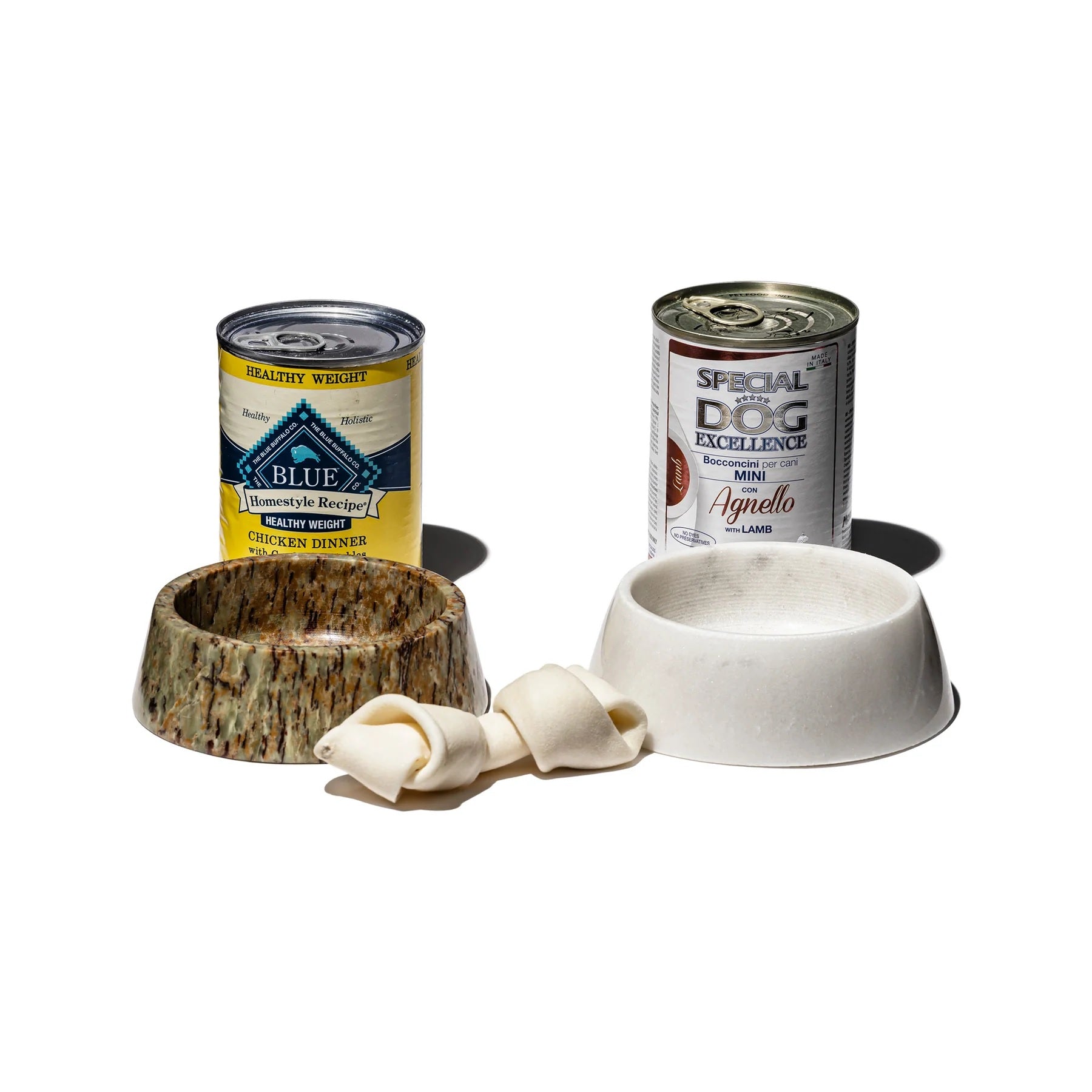 Two cans of dog food, a wooden bowl, a ceramic bowl, and a bone on a white background.