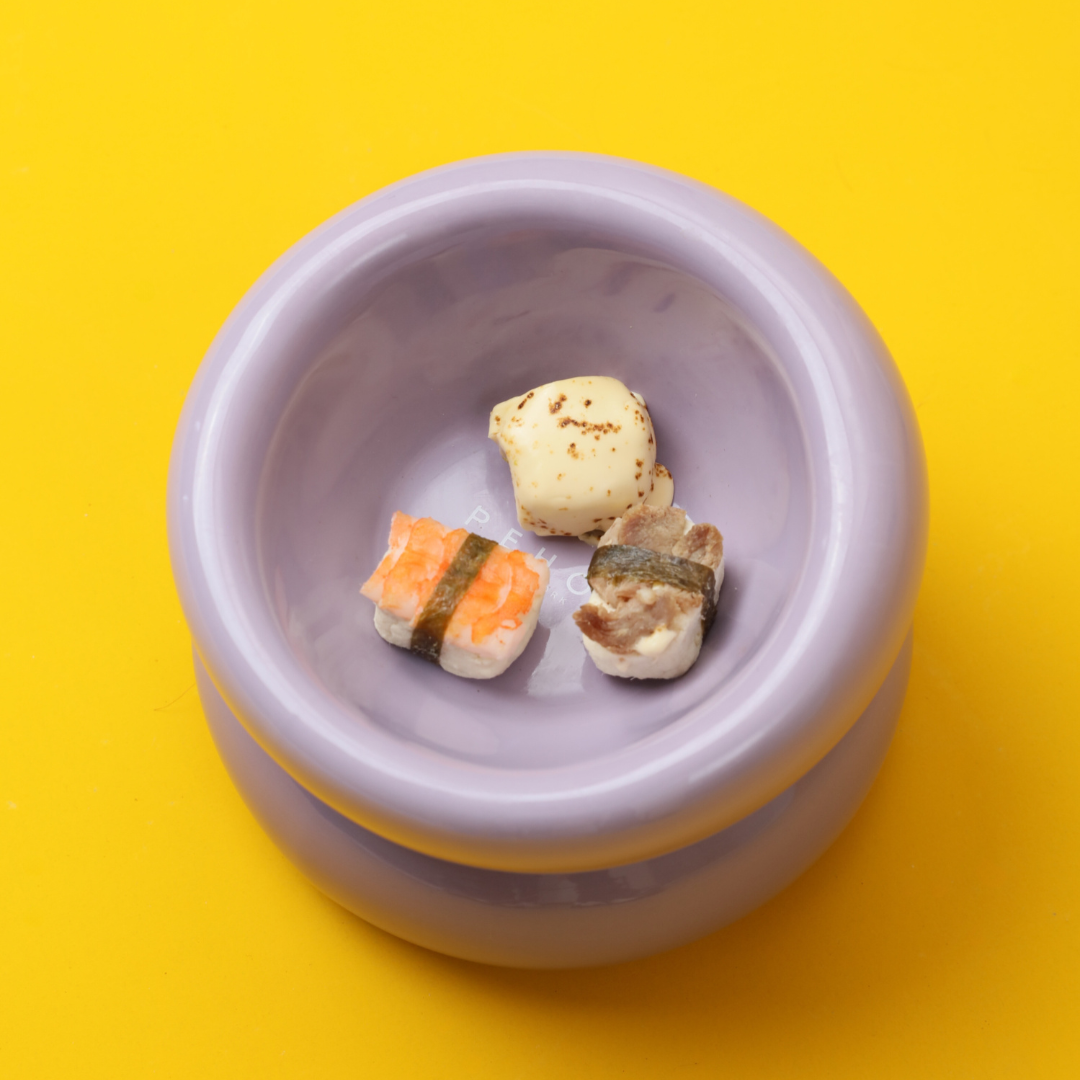 A light purple bowl contains three small sushi pieces, featuring rice, fish, and garnishes, against a yellow background.