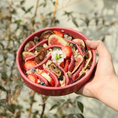A vibrant bowl of colorful fruit salad, including figs, strawberries, and cucumbers, topped with granola and a flower.
