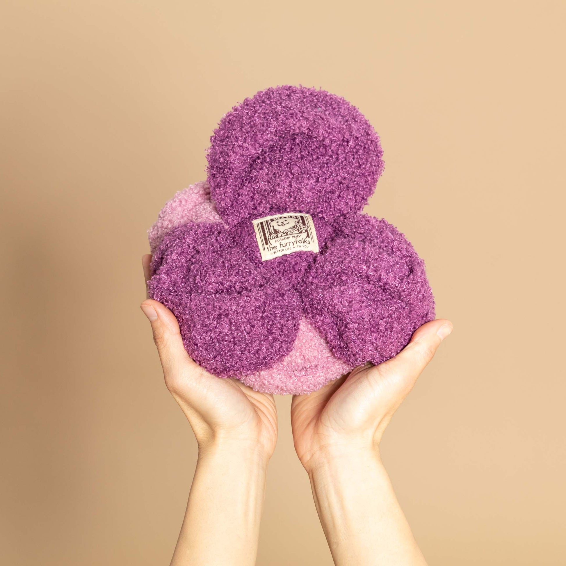 A pair of hands hold a fluffy, purple and pink cabbage toy, featuring a label in the center.