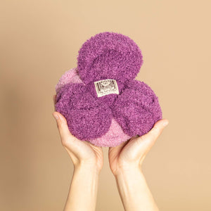 A pair of hands hold a fluffy, purple and pink cabbage toy, featuring a label in the center.