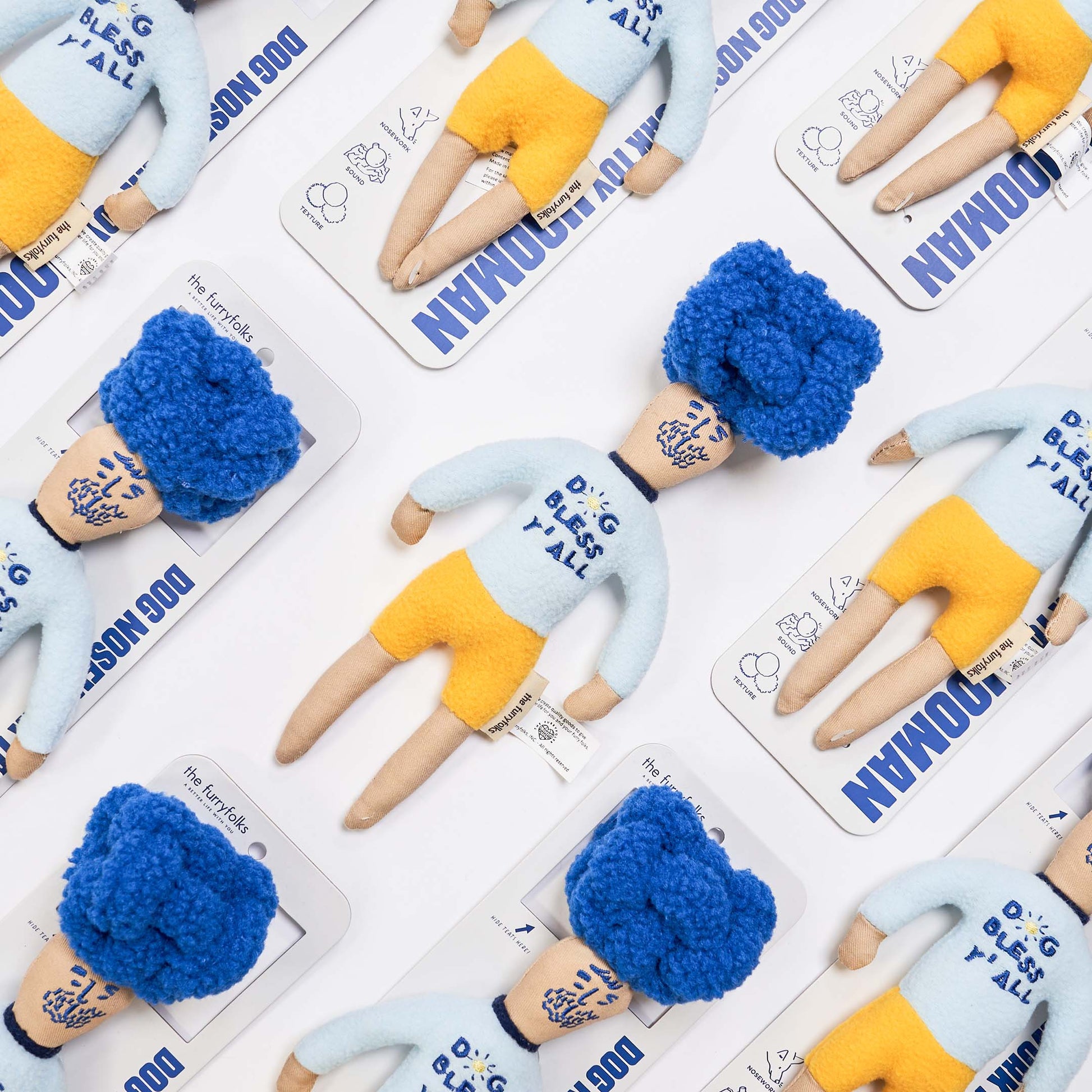 A flat lay of whimsical, handmade dolls with blue hair and yellow shorts, each on a branded backing card.
