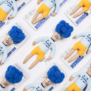 A flat lay of whimsical, handmade dolls with blue hair and yellow shorts, each on a branded backing card.
