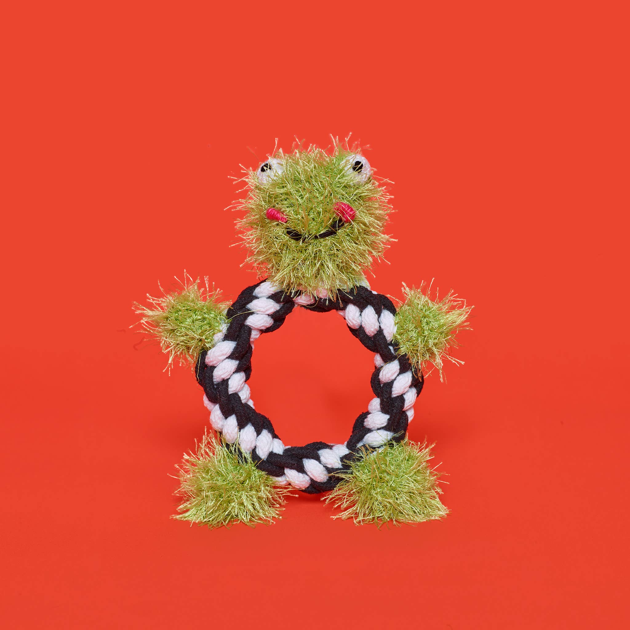 A cute pink rope toy shaped like a frog, with black and white rope for a body, set against a bright red background. 