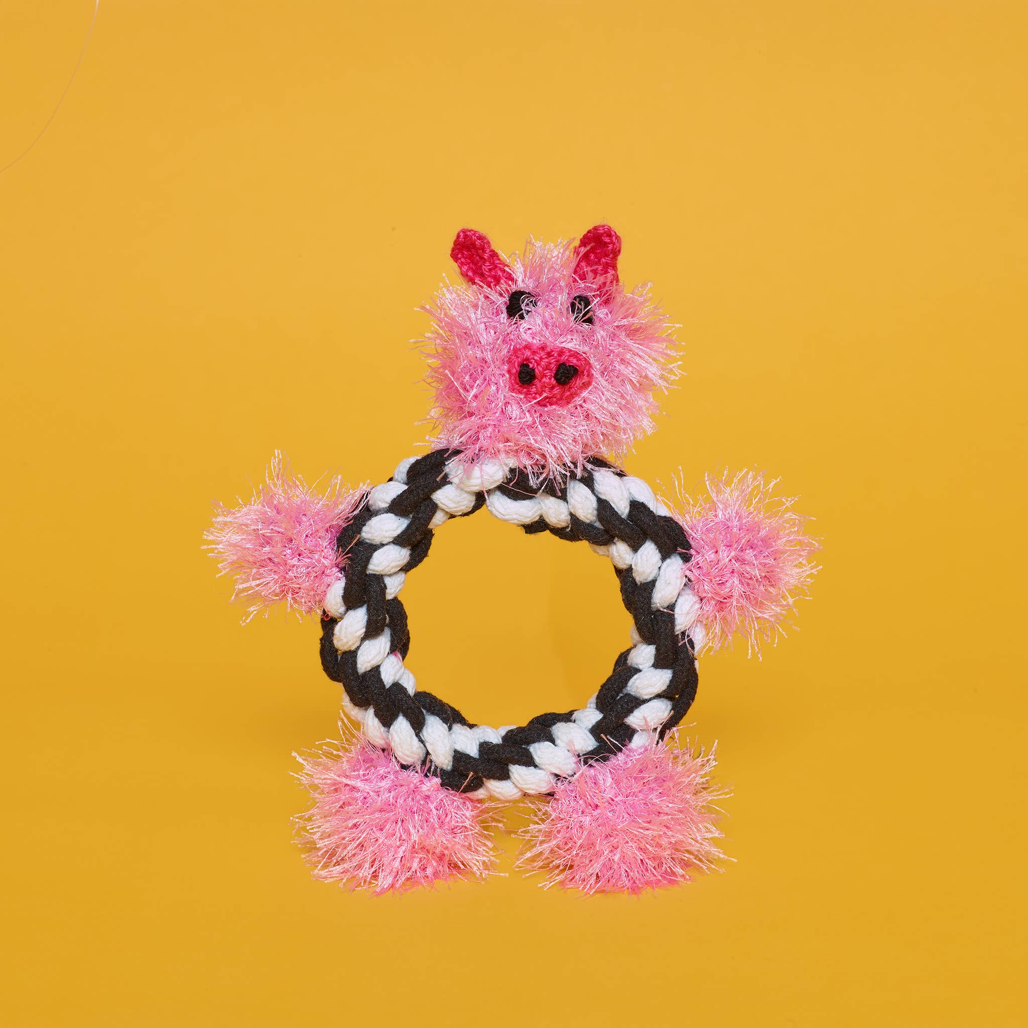 A cute pink rope toy shaped like a pig, with black and white rope for a body, set against a bright yellow background. 